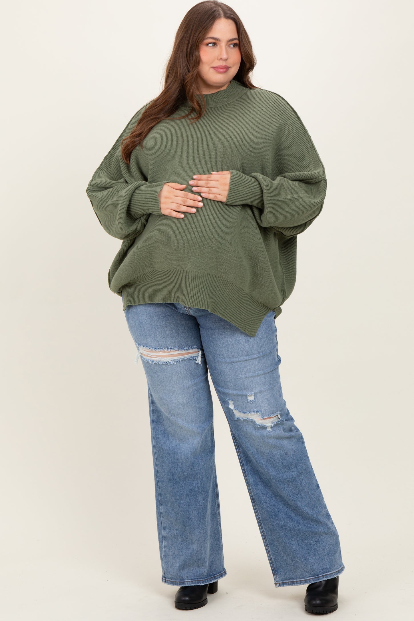 woman wearing an oversized olive green sweater and distressed blue jeans, showcasing her baby bump
