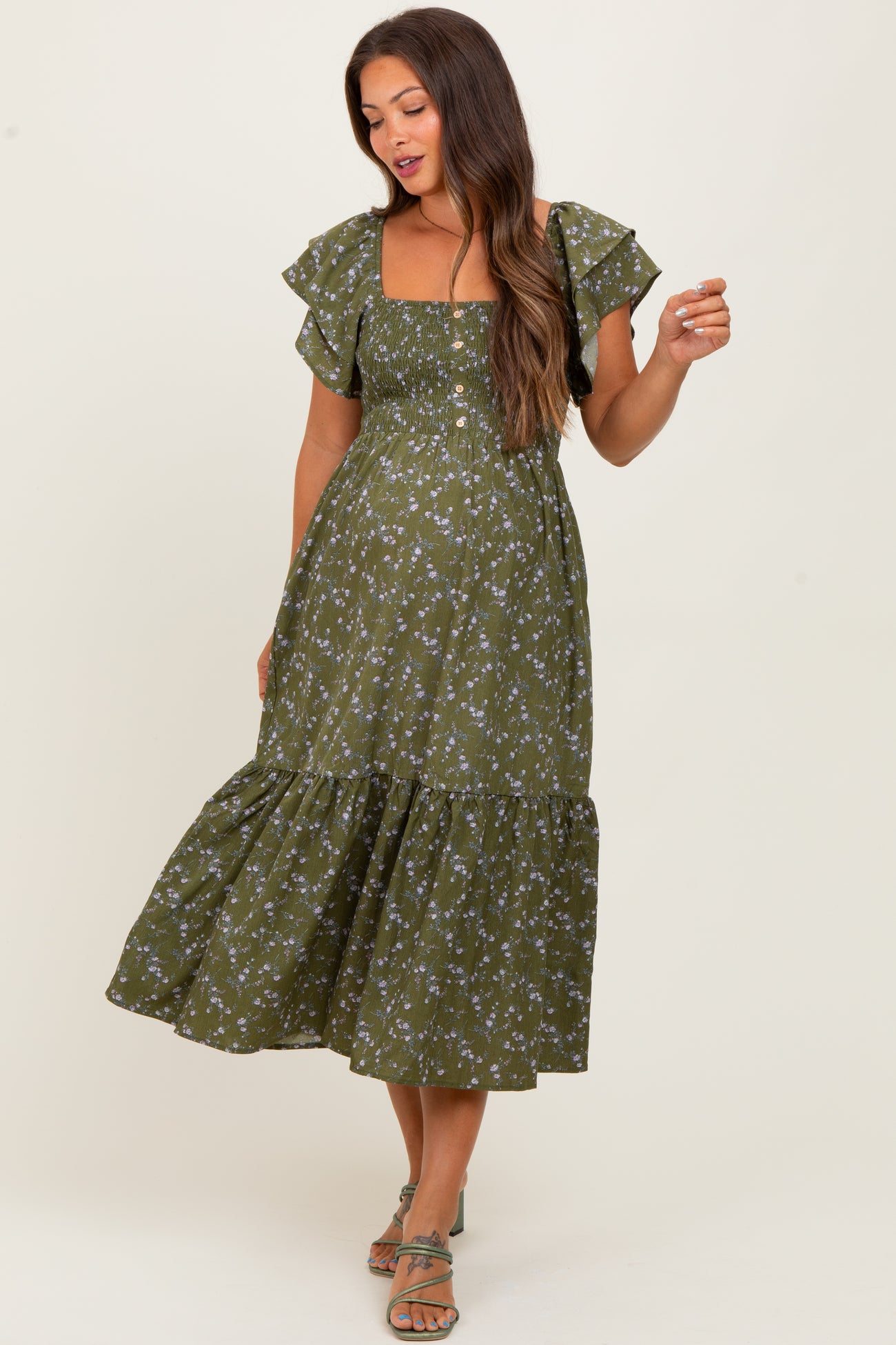 woman wearing a green floral print maternity dress with ruffled sleeves and a tiered skirt, holding one hand playfully