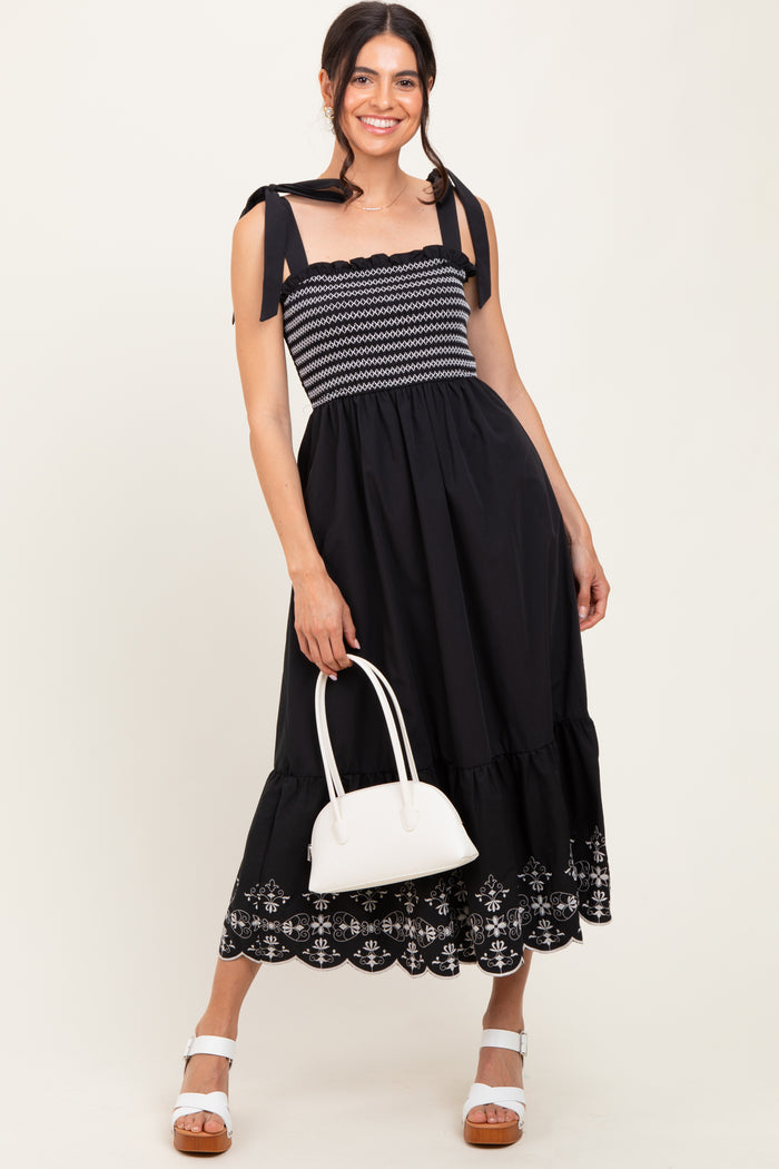 woman wearing a black elastic waist maxi dress with tie straps and embroidered hem, holding a white handbag