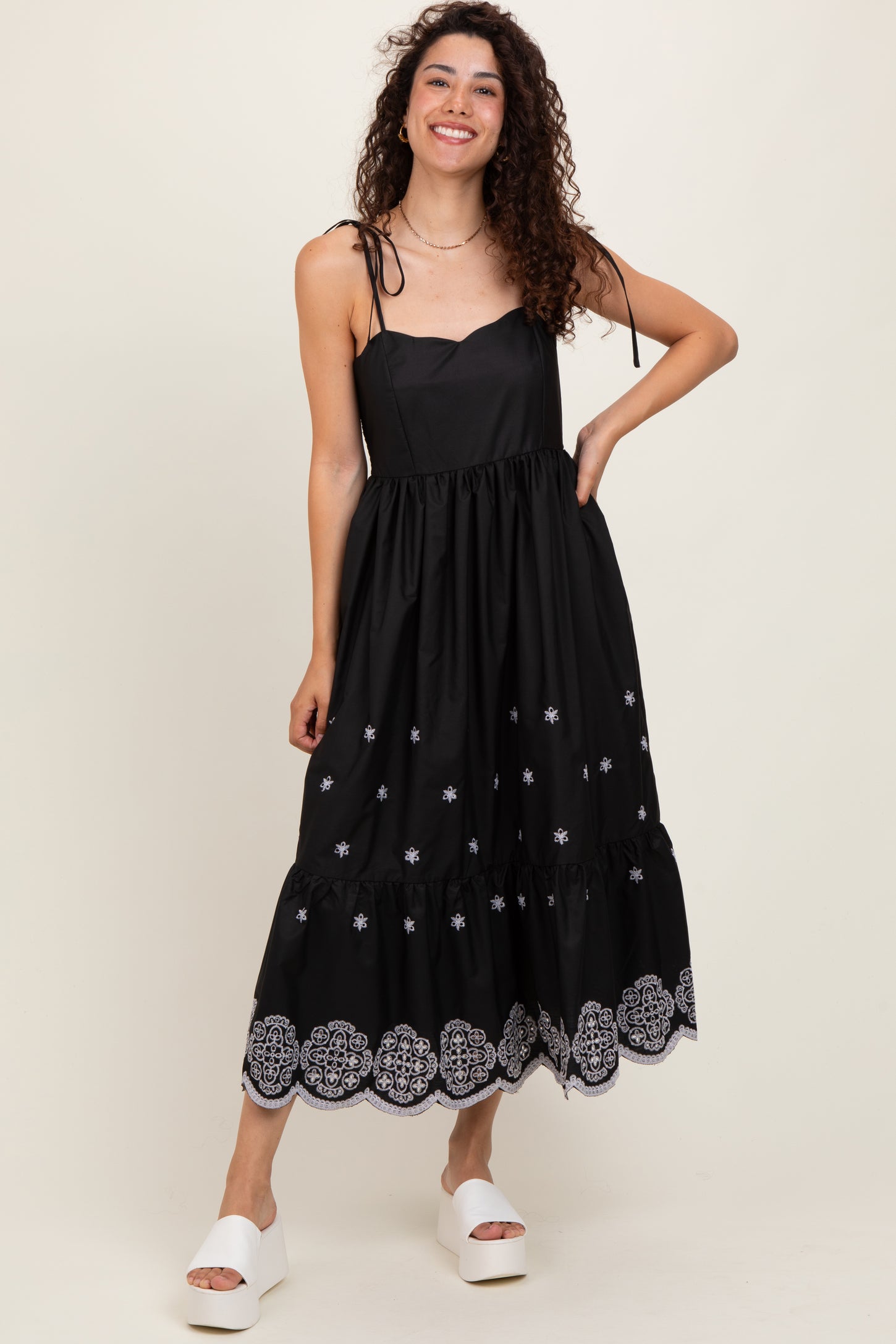 woman wearing a black sleeveless dress with embroidered details and a ruffle hem, standing in white platform sandals
