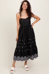 woman wearing a black sleeveless dress with embroidered details and a ruffle hem, standing in white platform sandals