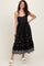 woman wearing a black sleeveless dress with embroidered details and a ruffle hem, standing in white platform sandals