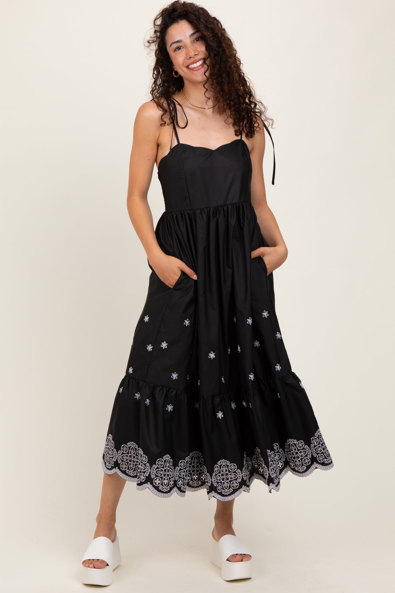 woman wearing a black midi dress with white embroidery and pockets, featuring adjustable straps and a tiered skirt.