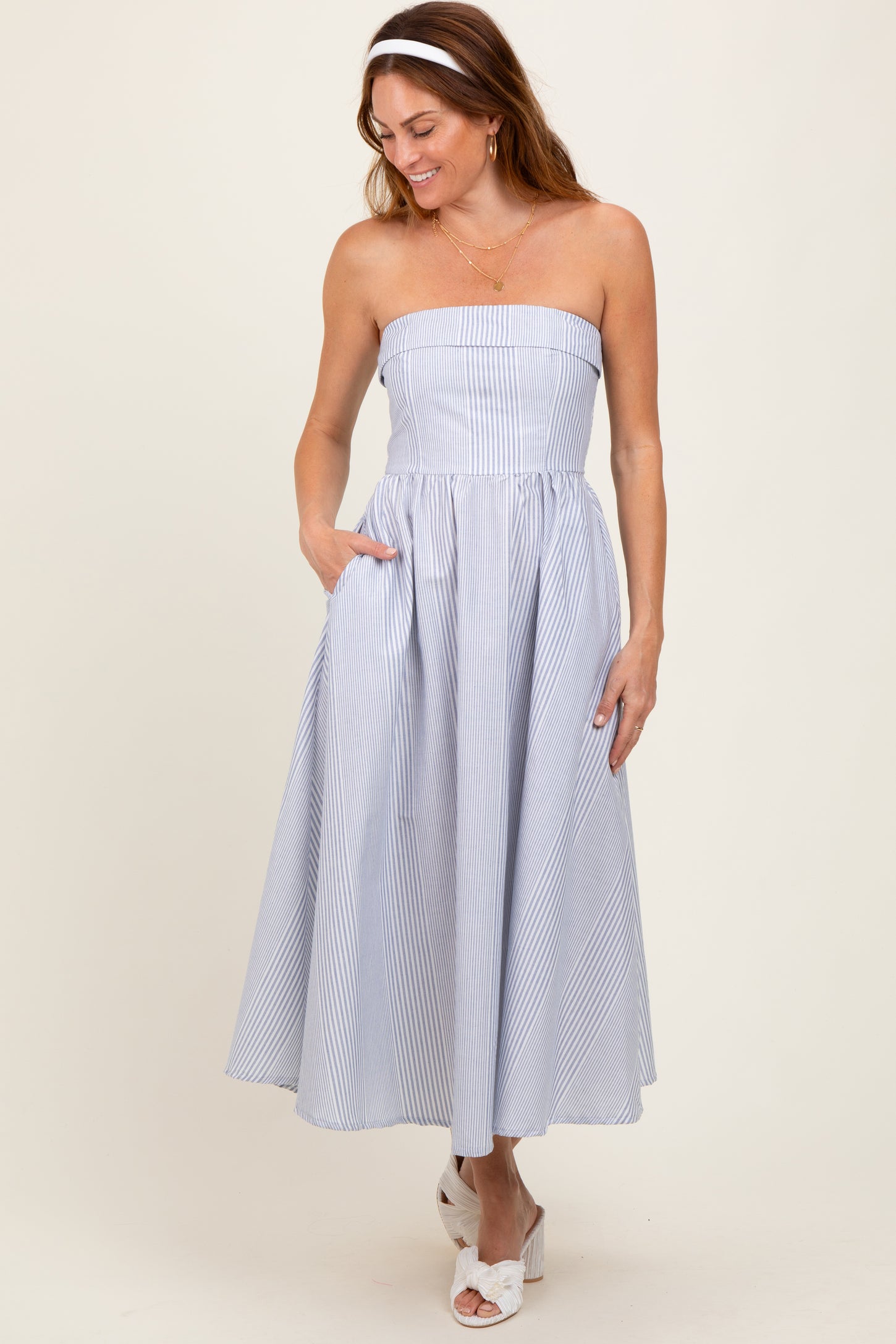 woman wearing a strapless blue and white striped midi dress with pockets, styled with a white headband and white sandals
