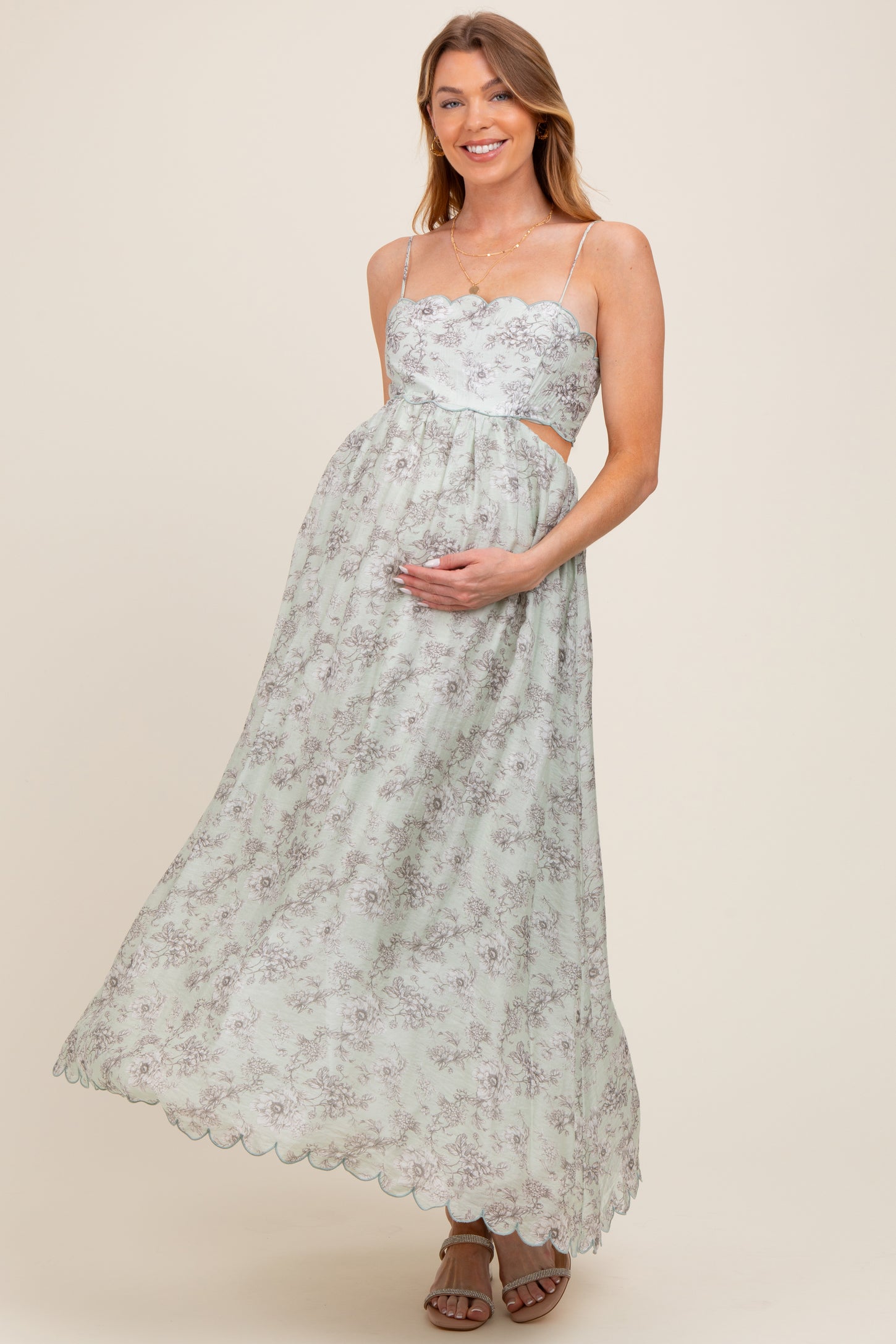woman wearing a light green floral maternity maxi dress with cutout sides and a scalloped hem, holding her baby bump