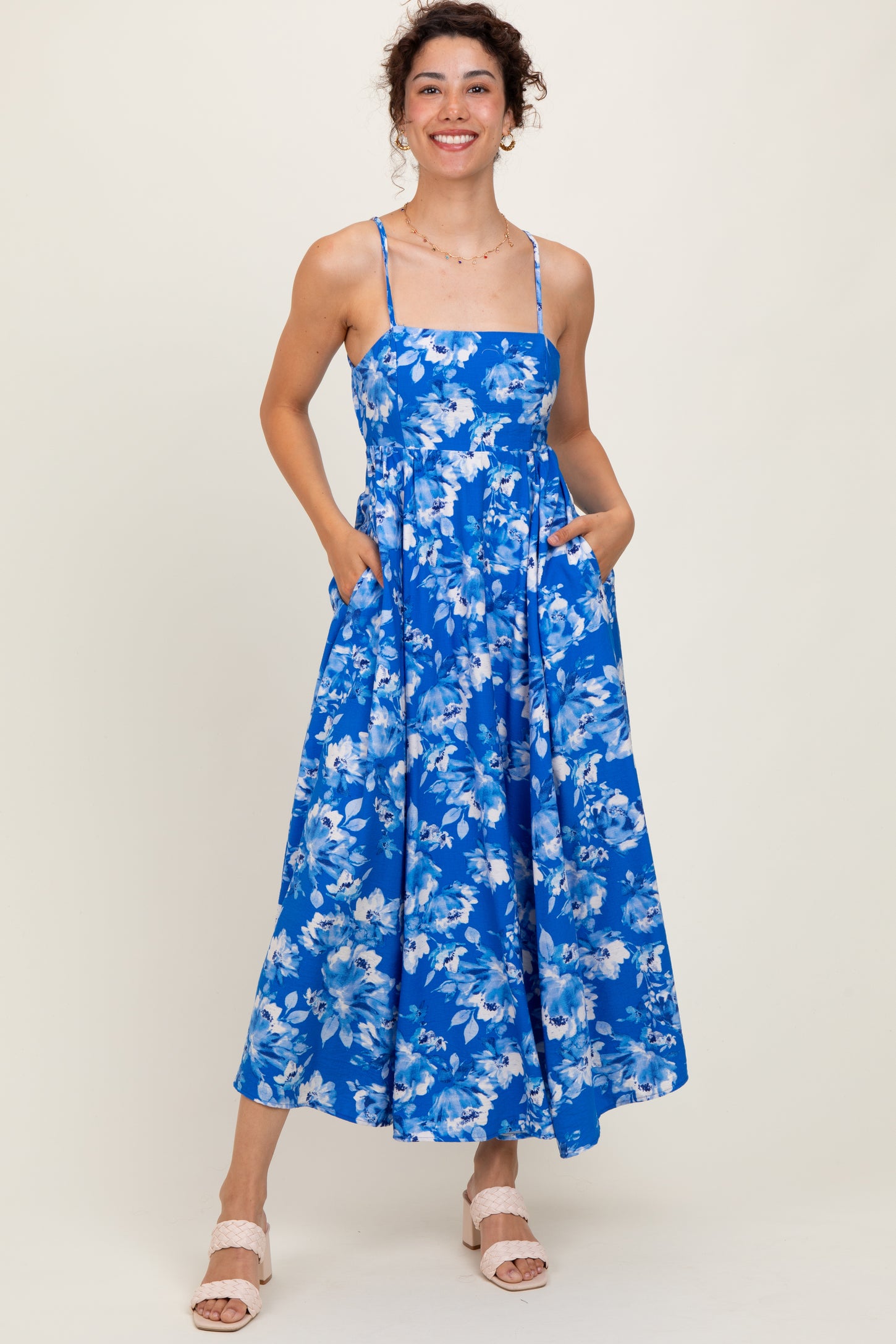 woman wearing a blue floral maxi dress with spaghetti straps and pockets, standing and smiling
