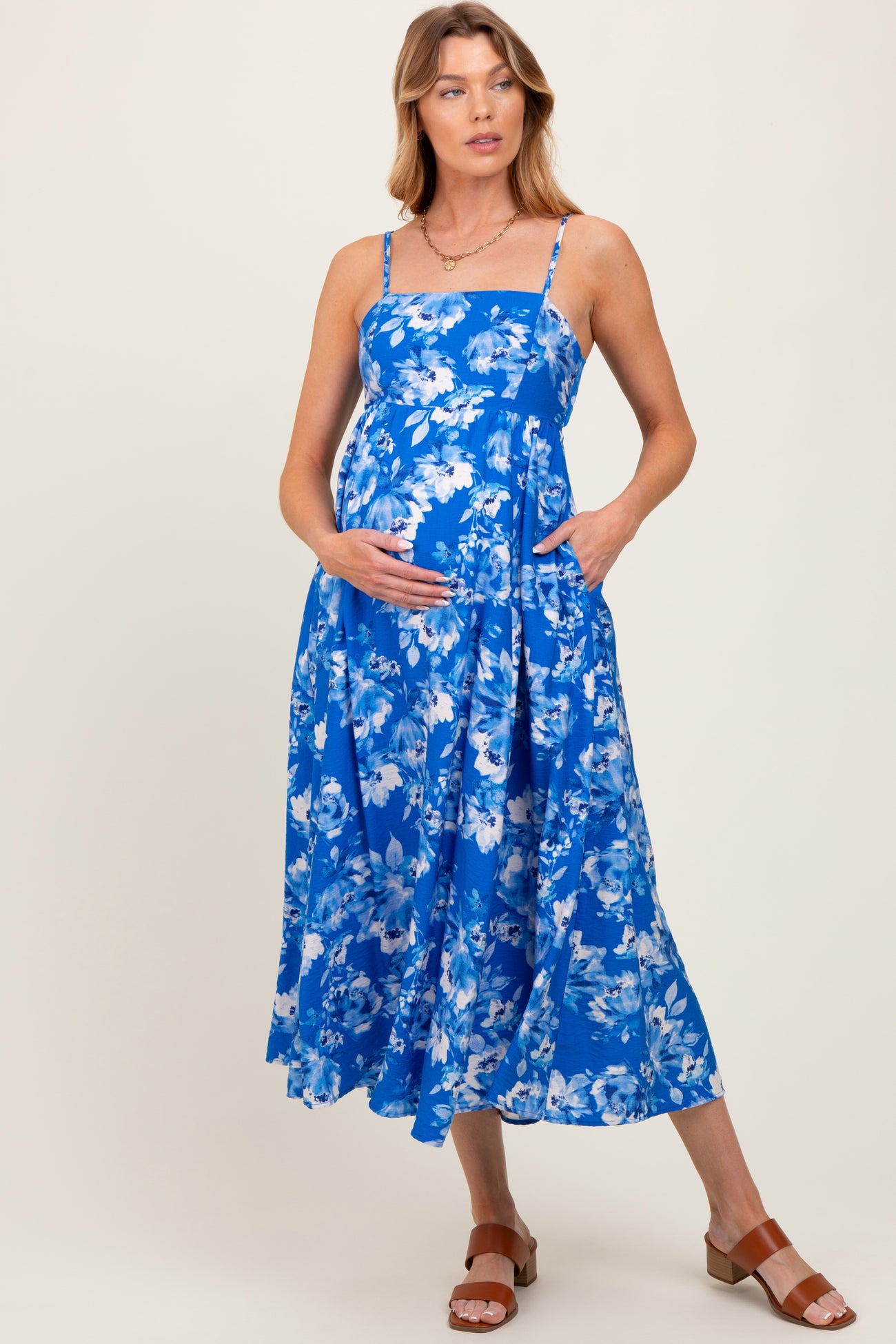 woman wearing a blue floral maternity dress with a fitted bodice, spaghetti straps, and a flared skirt, holding her baby bump