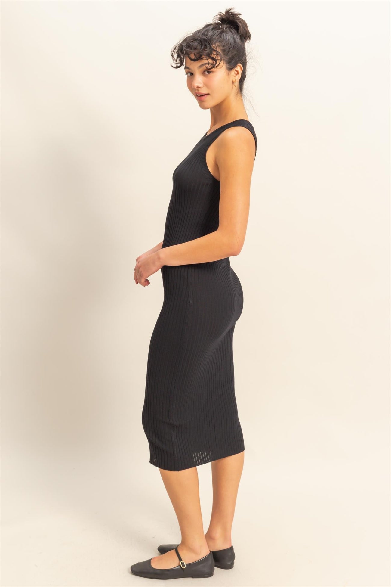woman wearing a fitted black ribbed midi dress with a sleeveless design and a side slit, standing in profile
