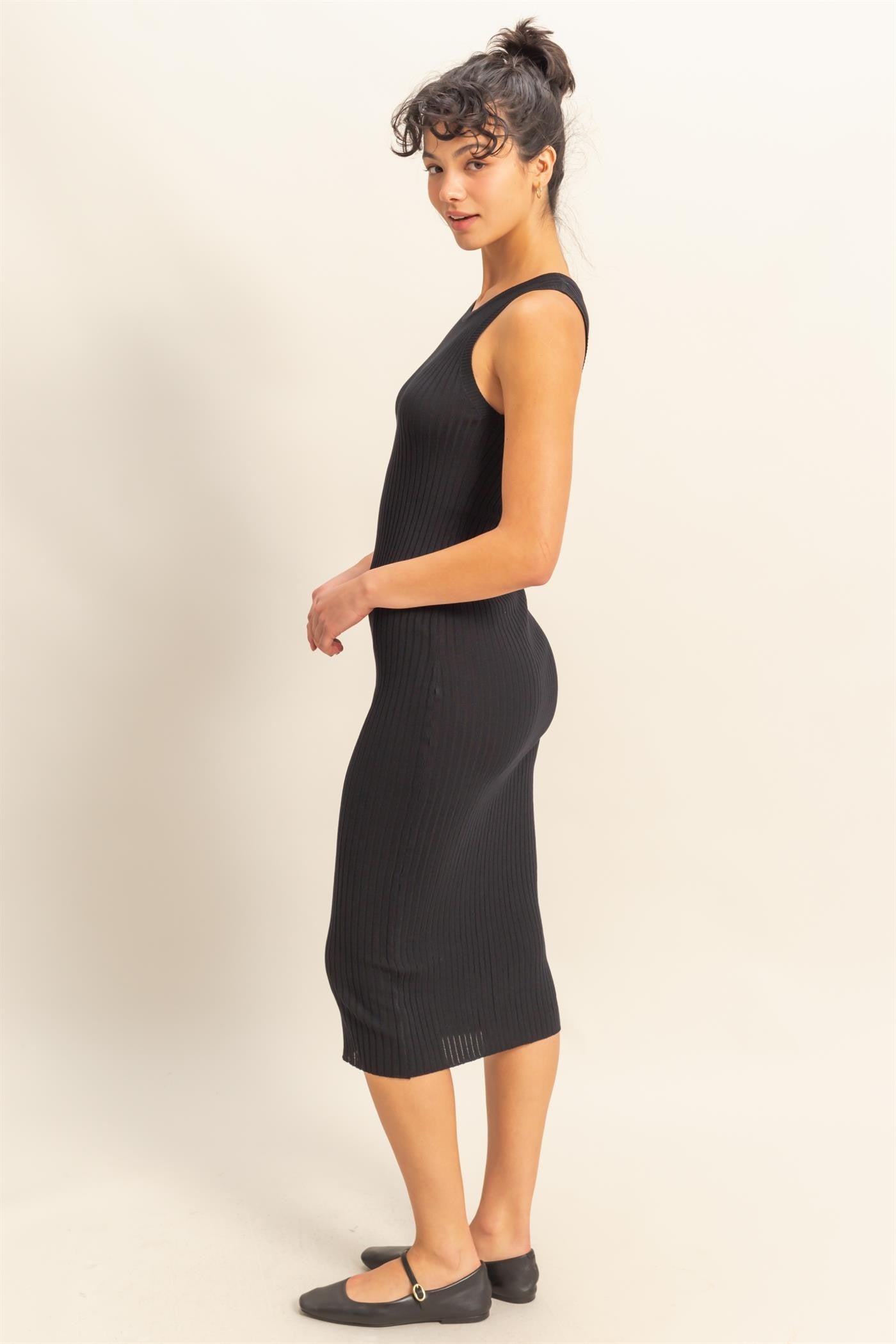 woman wearing a fitted black ribbed midi dress with a sleeveless design and a side slit, standing in profile