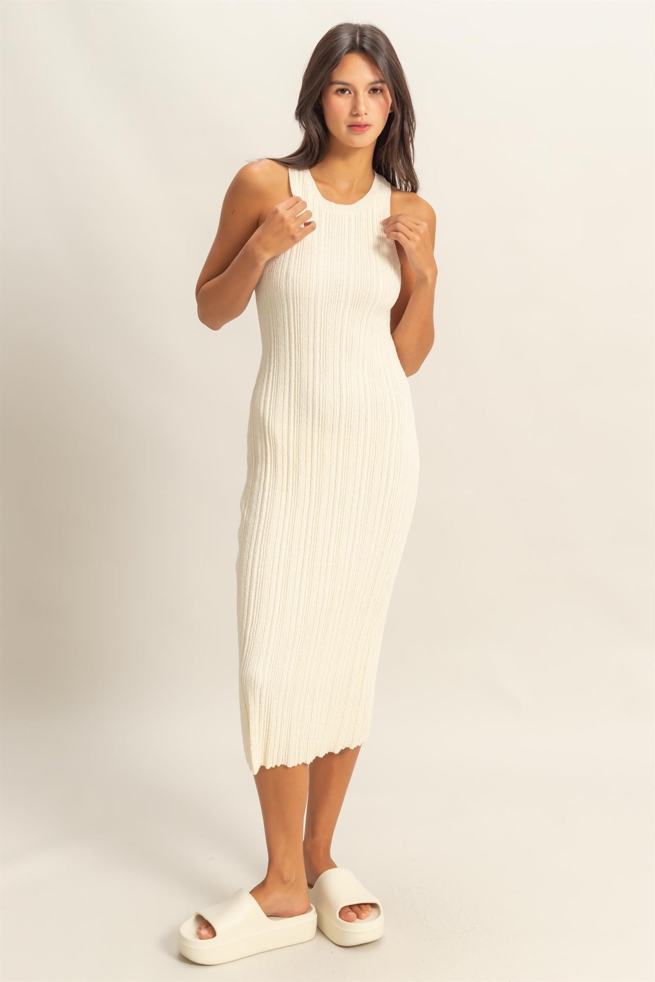 woman wearing a cream ribbed knit midi dress with a scoop neckline and side slits, paired with white slide sandals