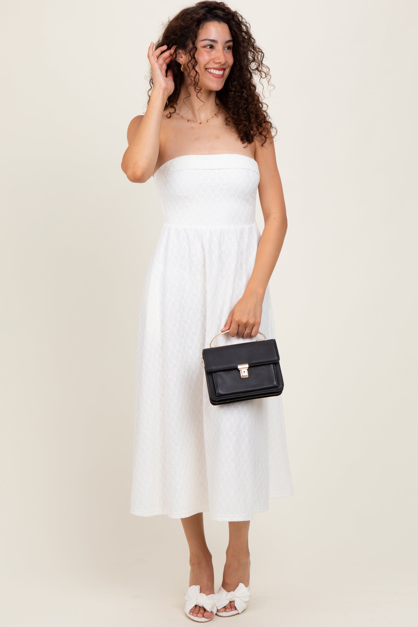 woman wearing a white strapless midi dress with a fitted bodice and flowy skirt, holding a black handbag