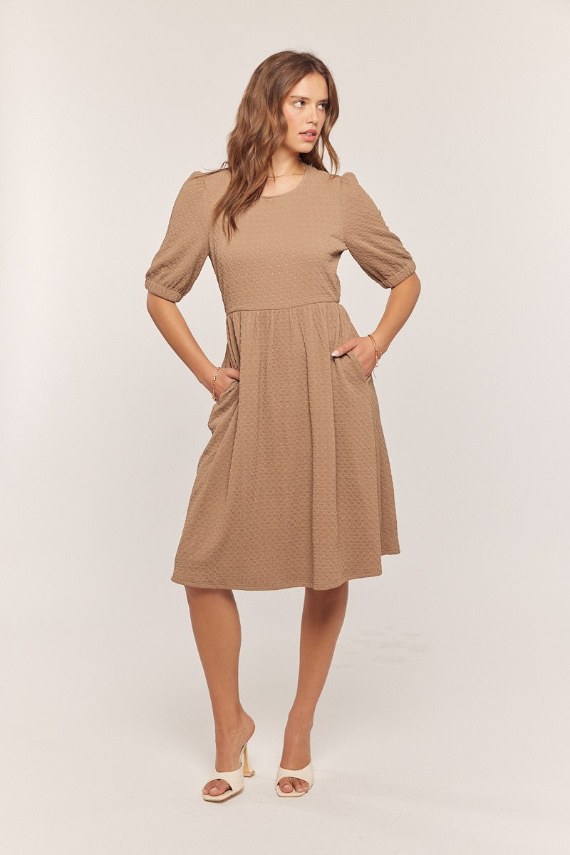 woman wearing a taupe puff sleeve dress with pockets and a fitted bodice, standing with one hand in pocket