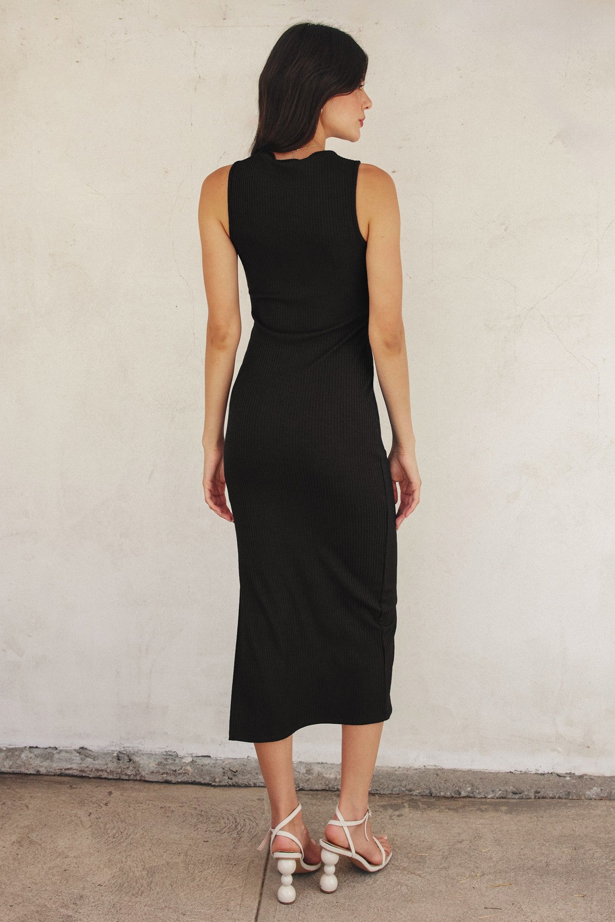 woman wearing a black ribbed midi dress with a fitted silhouette and back slit, paired with white strappy sandals