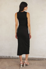 woman wearing a black ribbed midi dress with a fitted silhouette and back slit, paired with white strappy sandals
