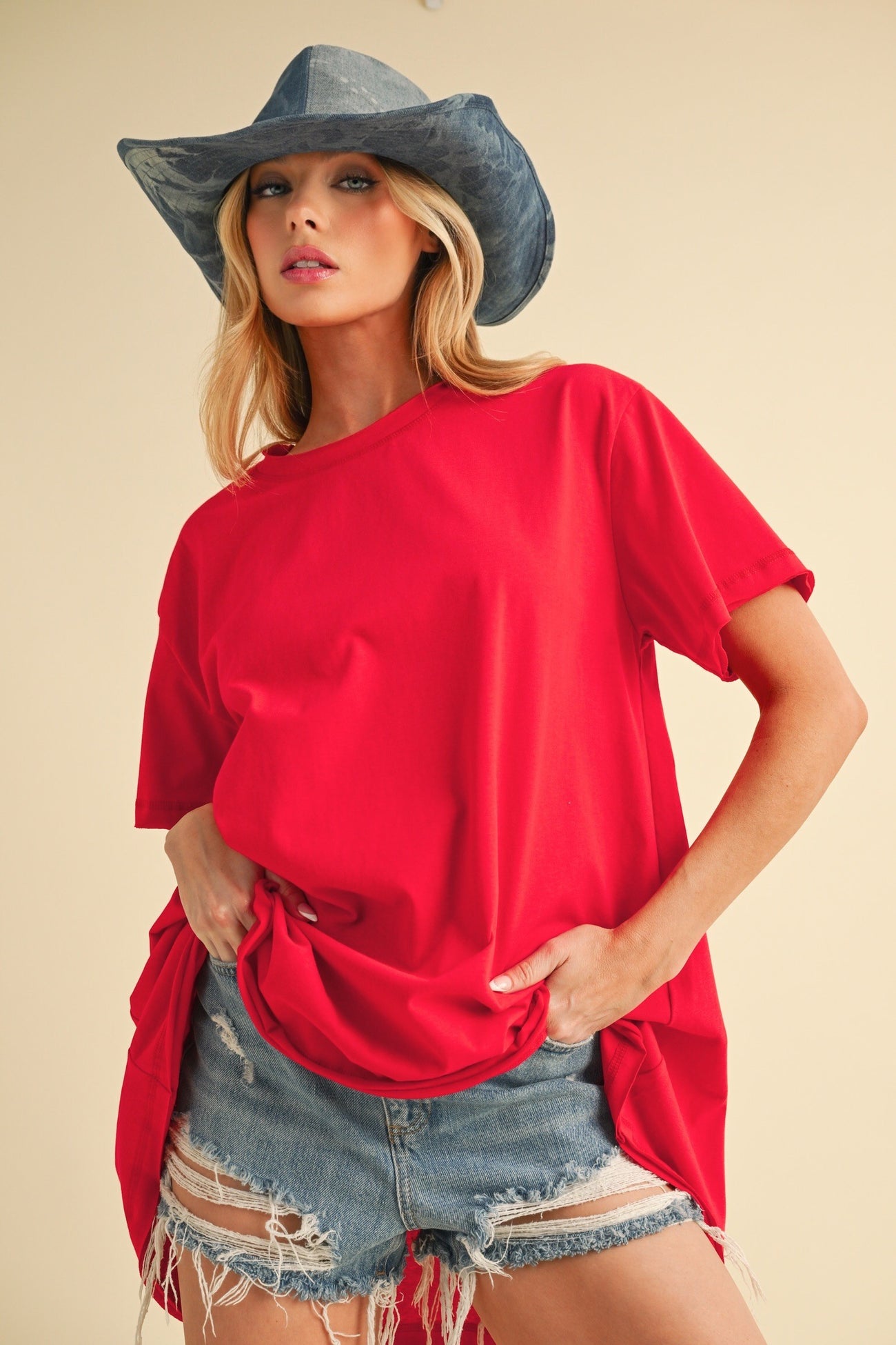 woman wearing a red oversized t-shirt and distressed denim shorts, accessorized with a blue cowboy hat