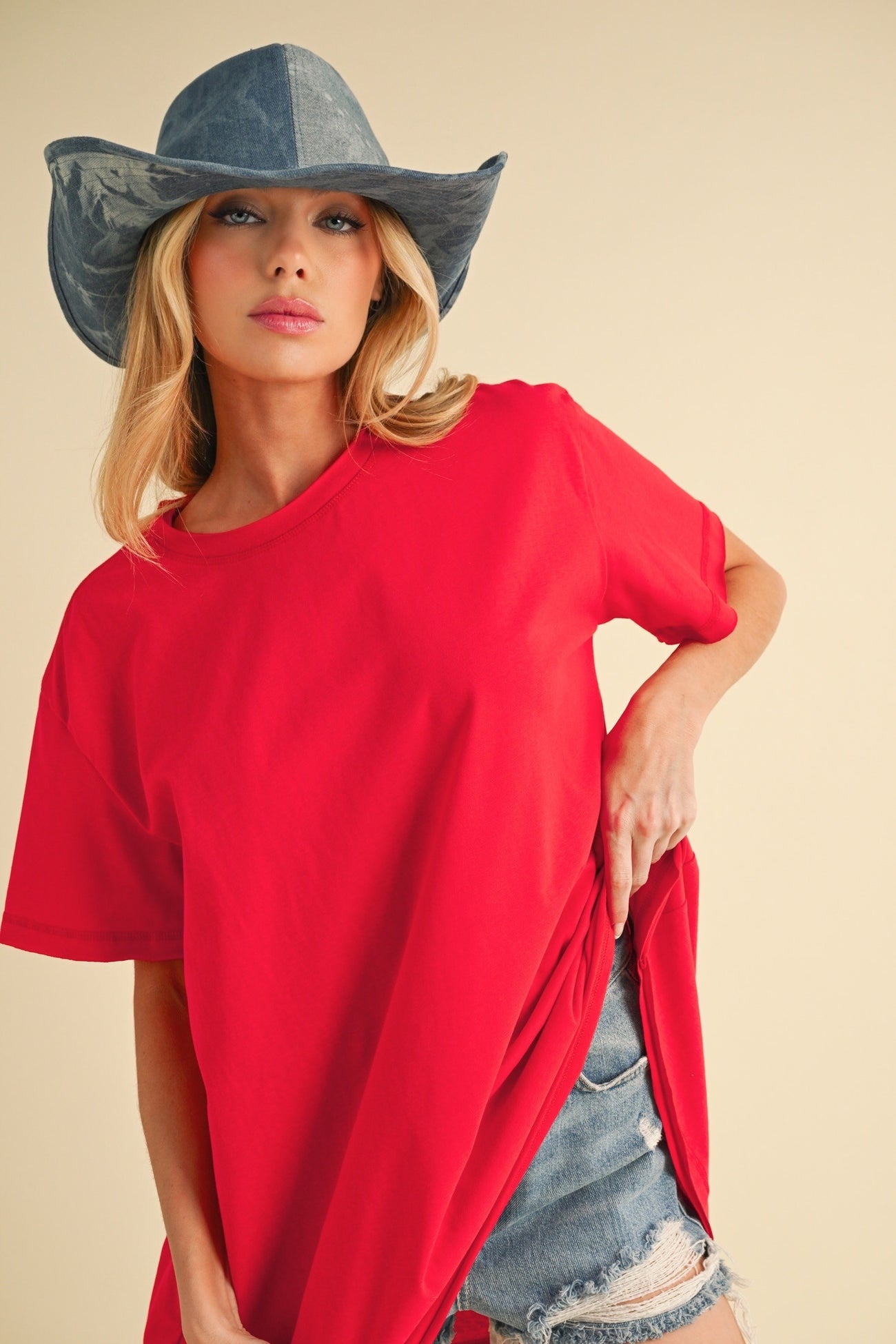 woman wearing a bright red oversized t-shirt with distressed denim shorts and a light blue wide-brimmed hat