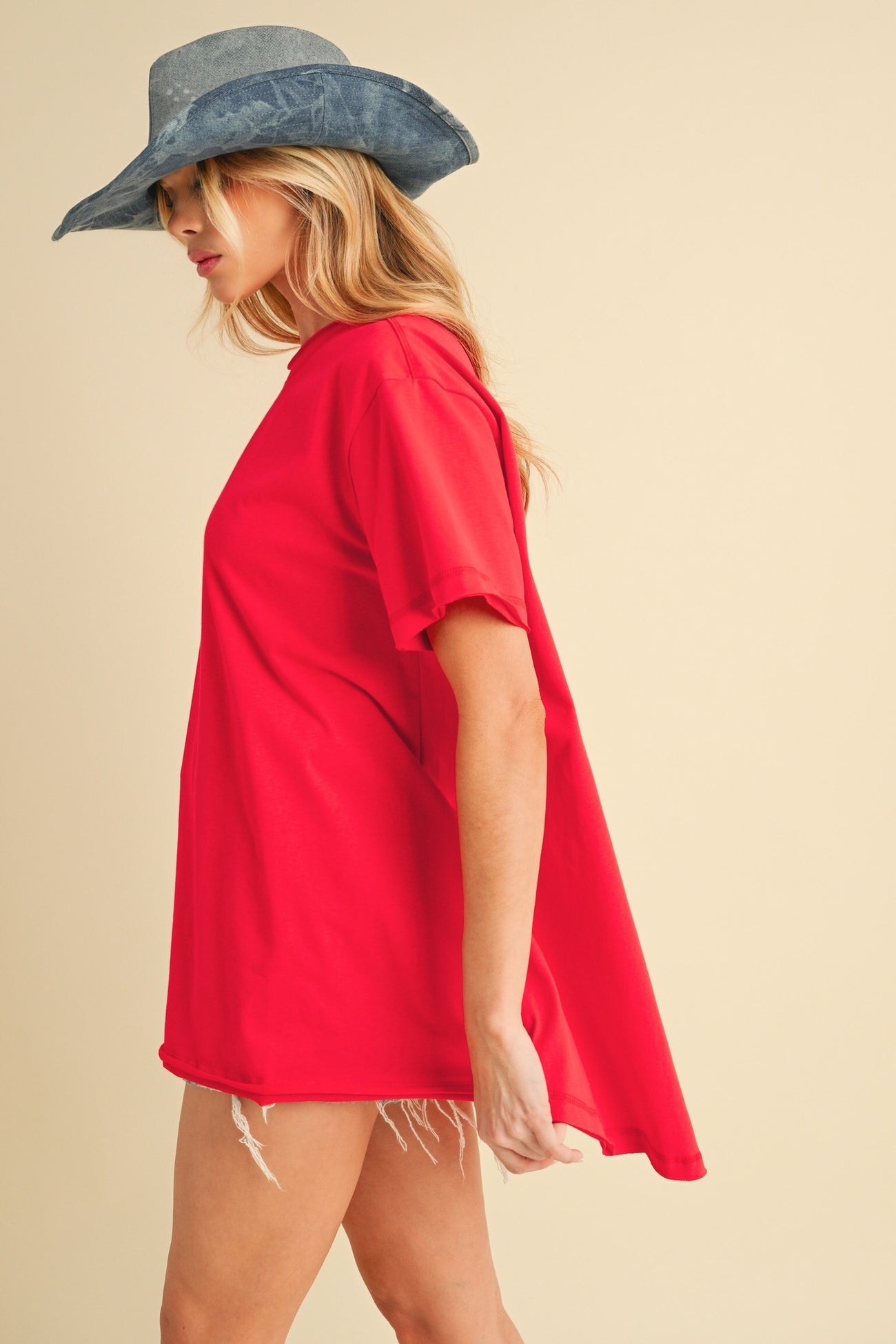 woman wearing a red oversized t-shirt and a gray wide-brim hat, standing against a light background