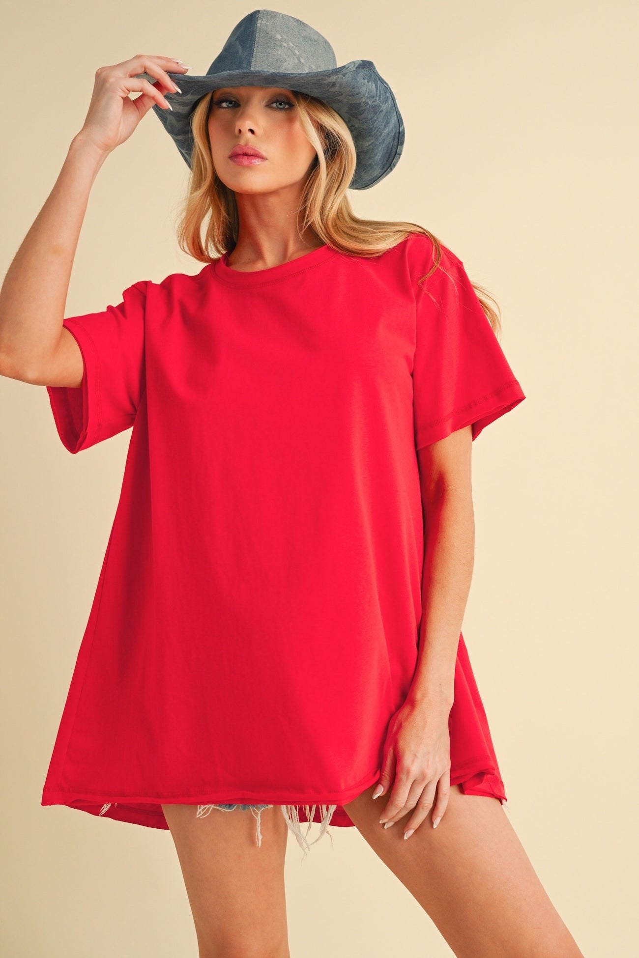 woman wearing a bright red oversized t-shirt with rolled sleeves and distressed denim shorts, accessorized with a blue hat