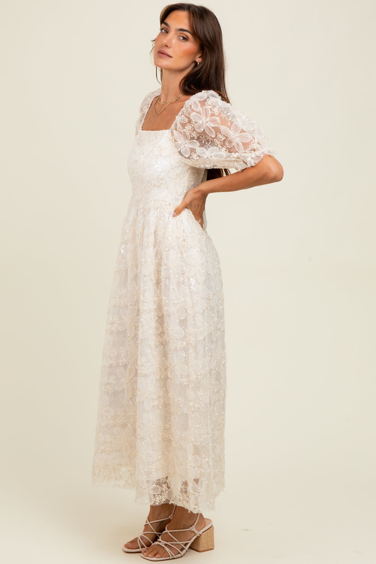 woman wearing a cream floral lace maxi dress with puff sleeves and a square neckline, standing with one hand on her hip