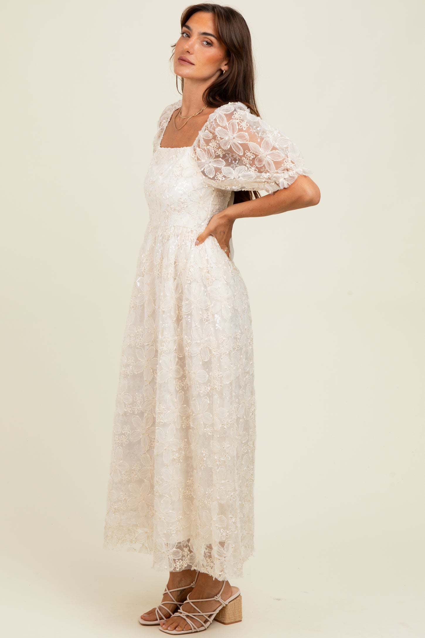woman wearing a cream floral lace maxi dress with puff sleeves and a square neckline, standing with one hand on her hip
