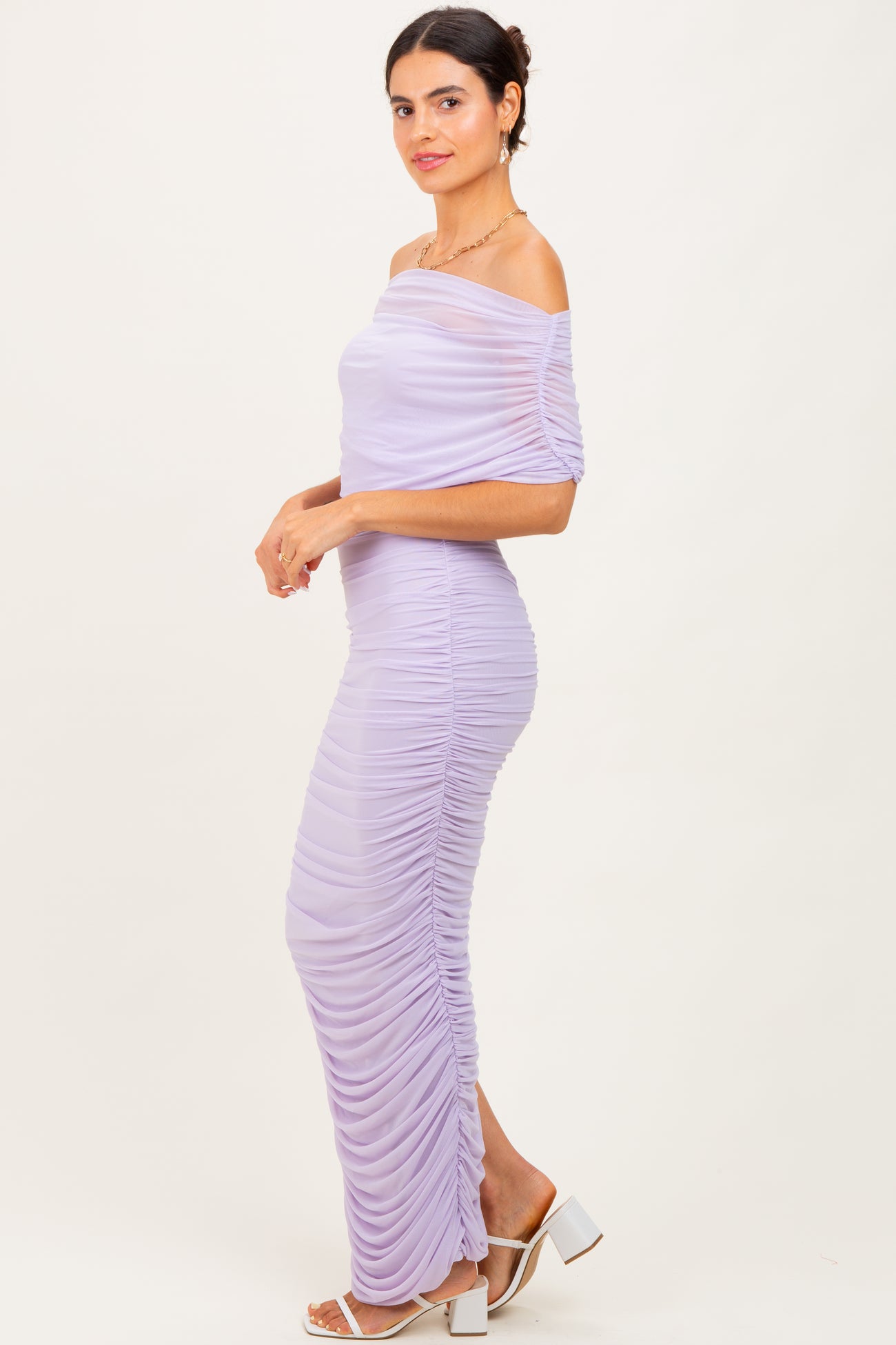 woman wearing a lilac off-the-shoulder ruched maxi dress with a fitted silhouette and side slit, standing sideways