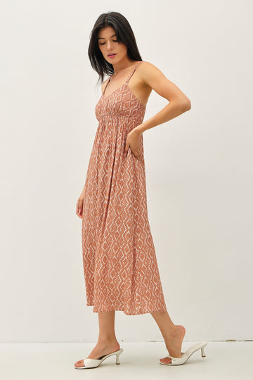 woman wearing a rust colored maxi dress with a geometric pattern and spaghetti straps, standing with one hand in pocket