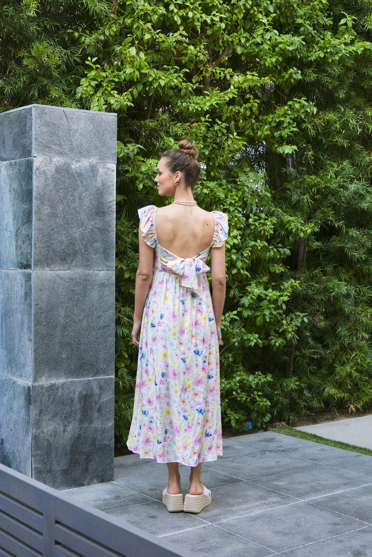 woman wearing a floral midi dress with a back tie detail and flutter sleeves, standing outdoors against greenery