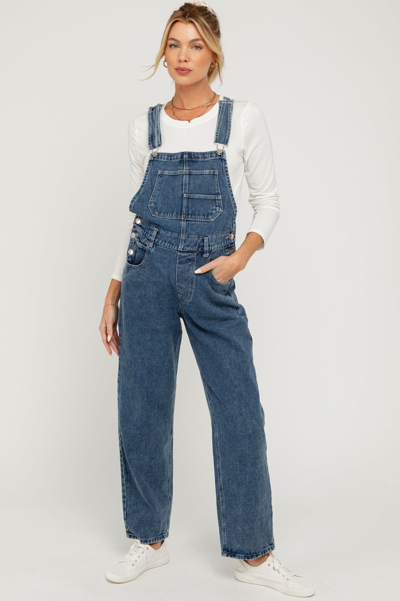 woman wearing a pair of blue denim maternity overalls with a white long sleeve top and sneakers