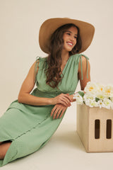 woman wearing a green polka dot midi dress with tie straps, smiling and seated next to a wooden box of flowers