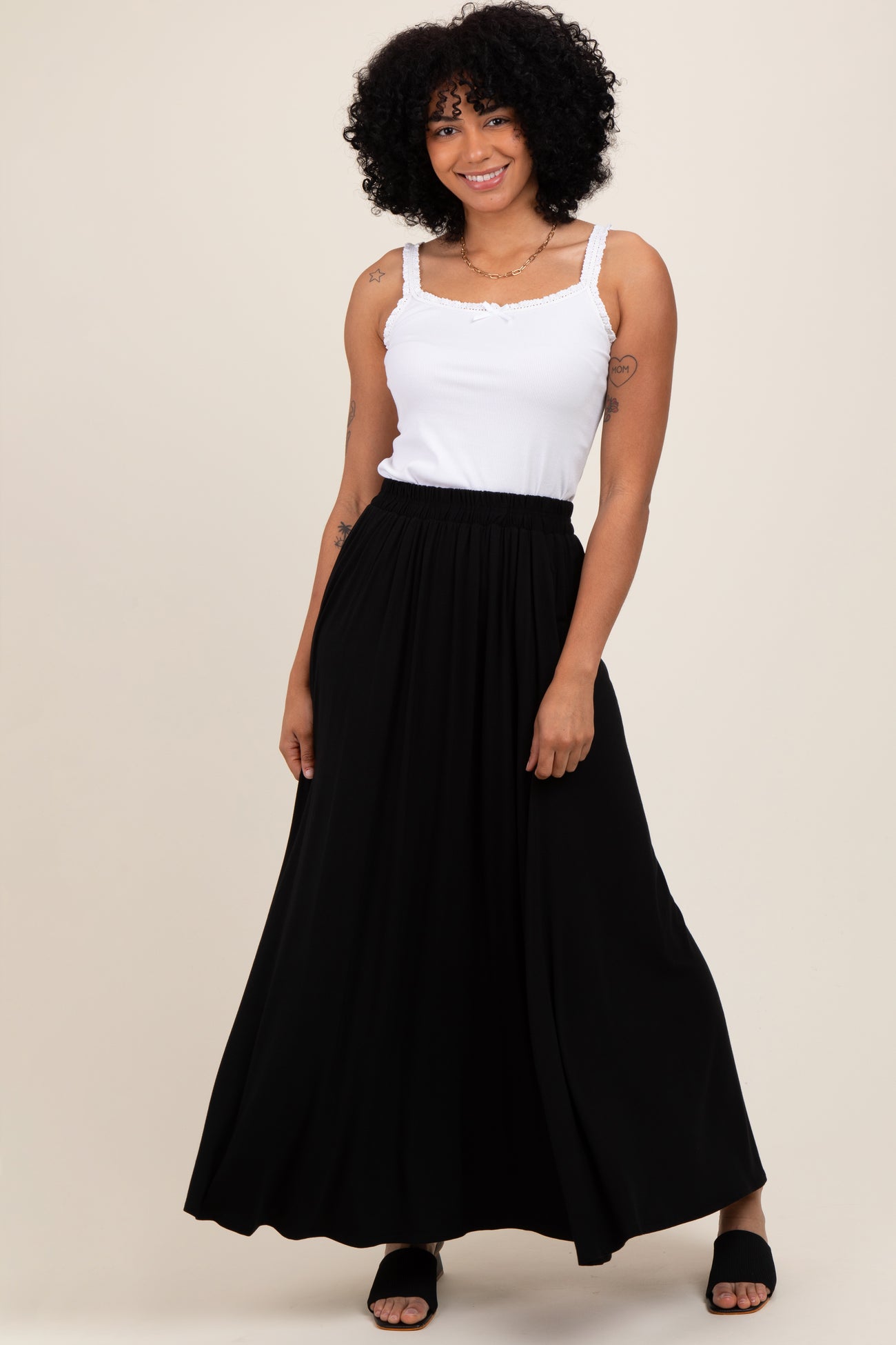woman wearing a white tank top and a black maxi skirt, standing with a smile against a neutral background
