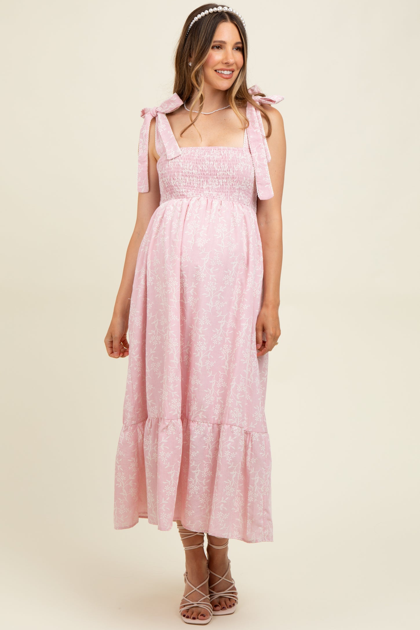 woman wearing a light pink floral maternity dress with bow straps and a flared skirt, showing her baby bump