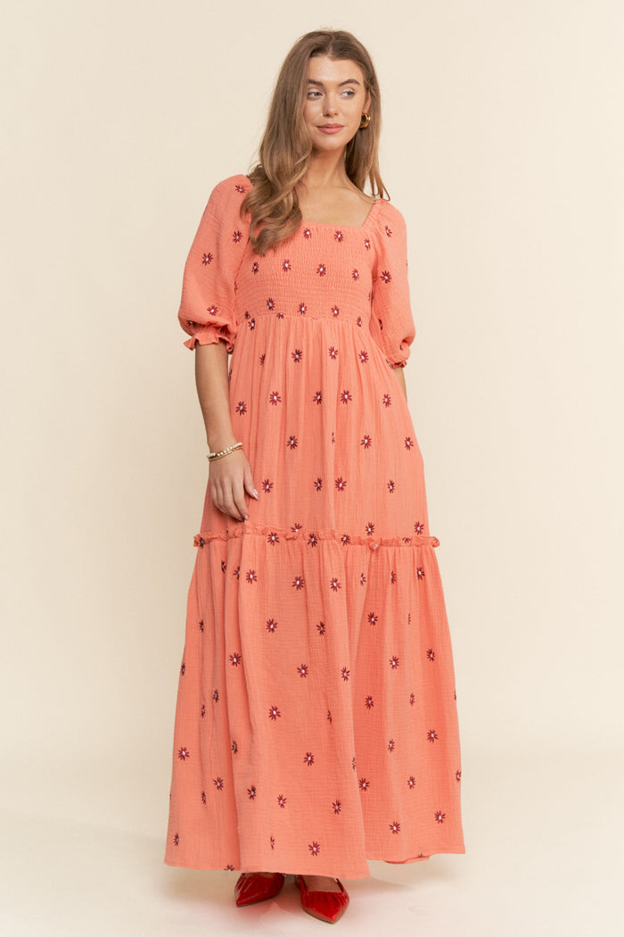woman wearing a coral maxi dress with puff sleeves and floral embroidery, standing against a neutral background