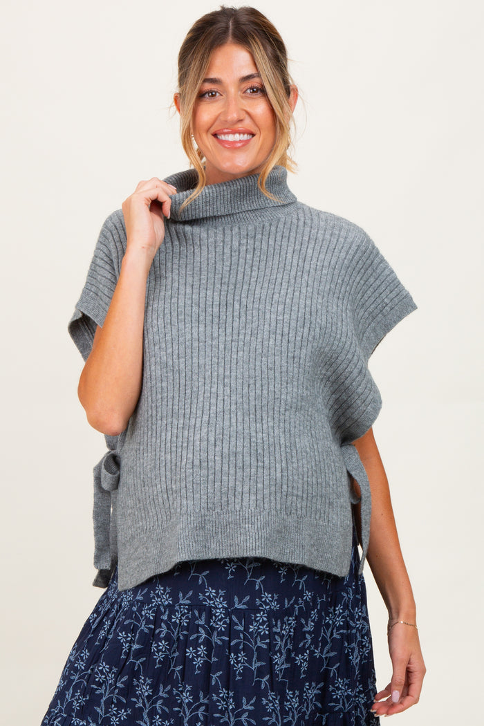 woman wearing a gray ribbed maternity poncho top with side ties and a blue floral print skirt, smiling and holding the neckline