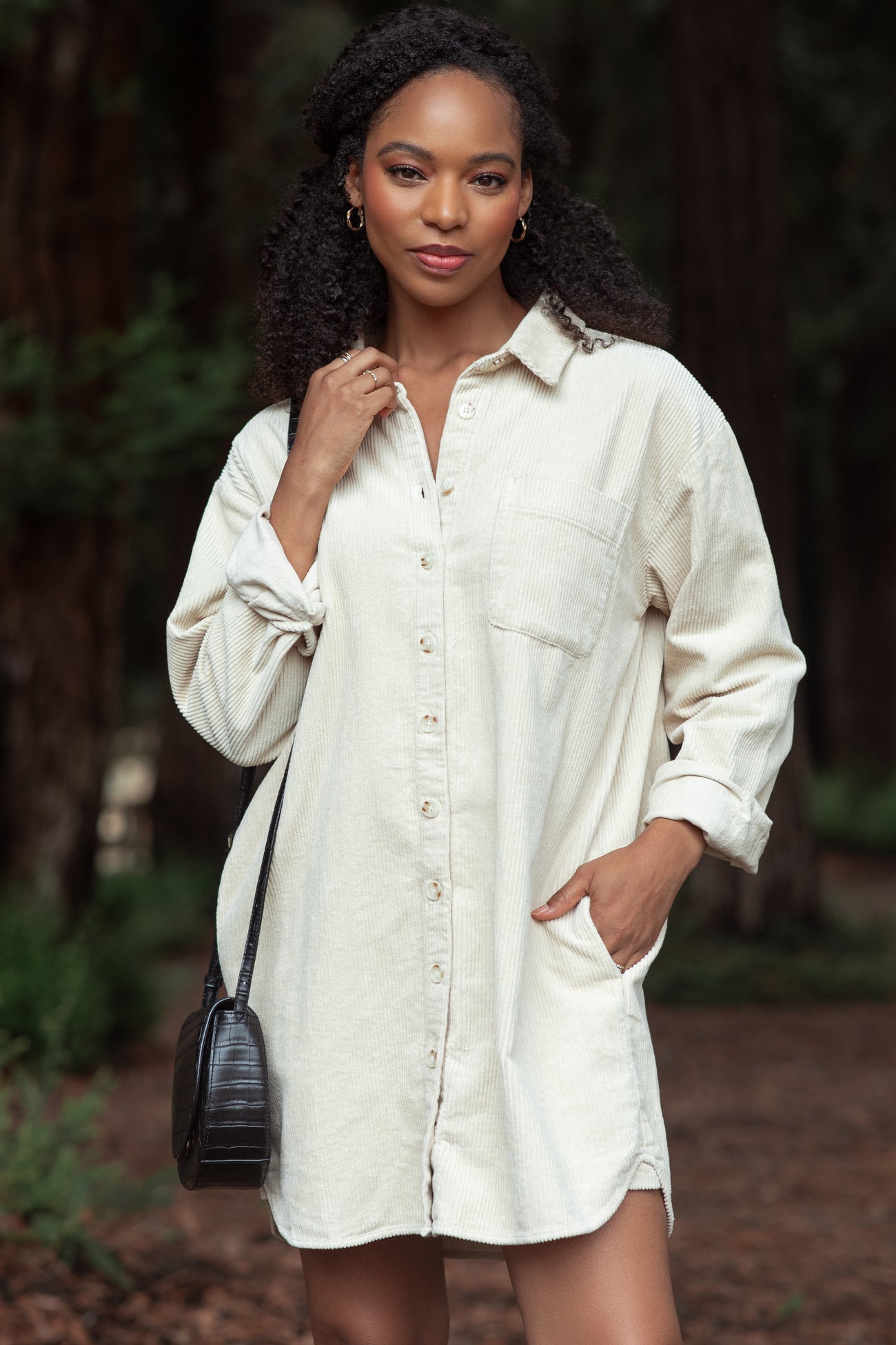 woman wearing a beige corduroy shirt dress with a button front, long sleeves, and a side pocket, standing outdoors