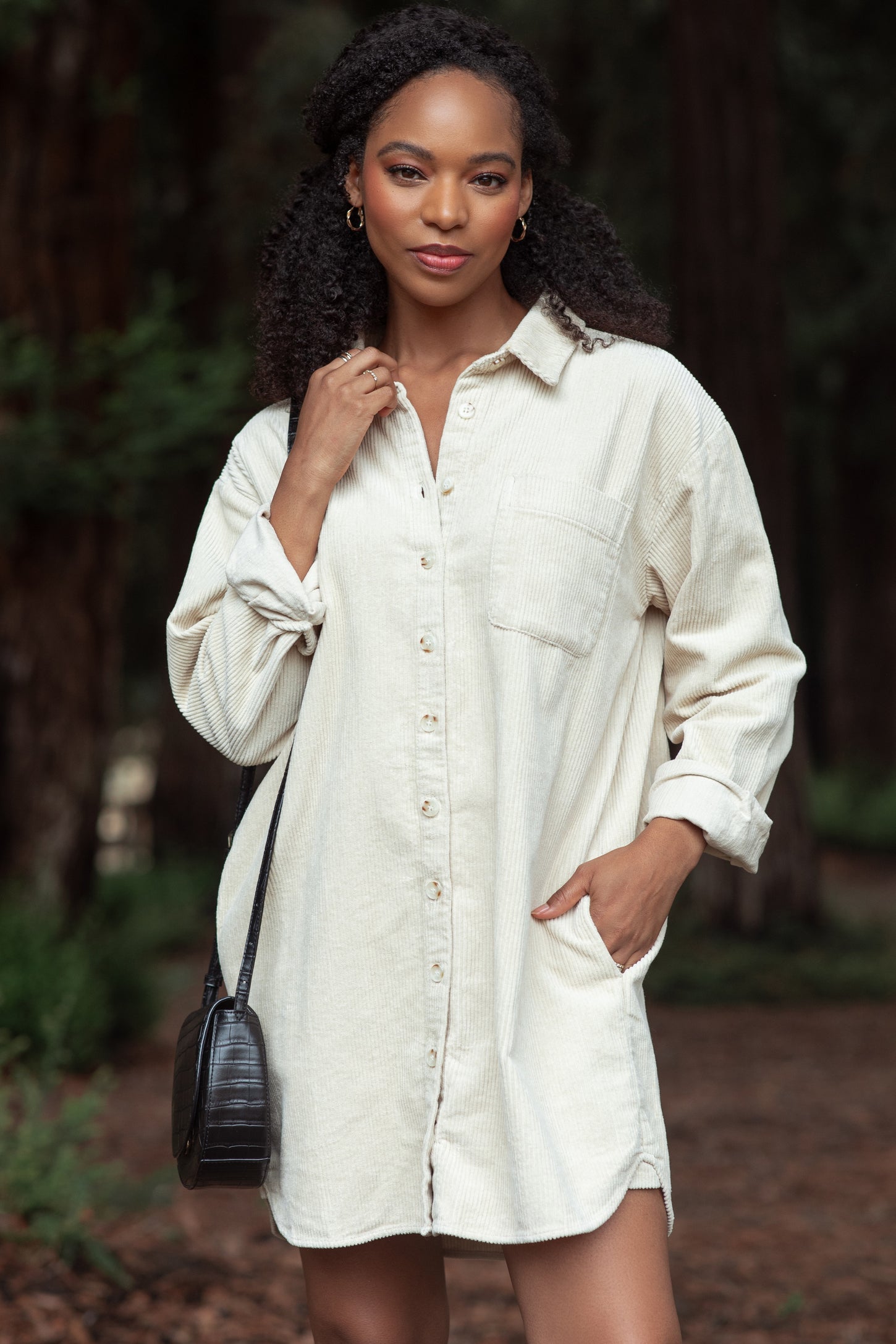 woman wearing a beige corduroy shirt dress with a button front, long sleeves, and a side pocket, standing outdoors