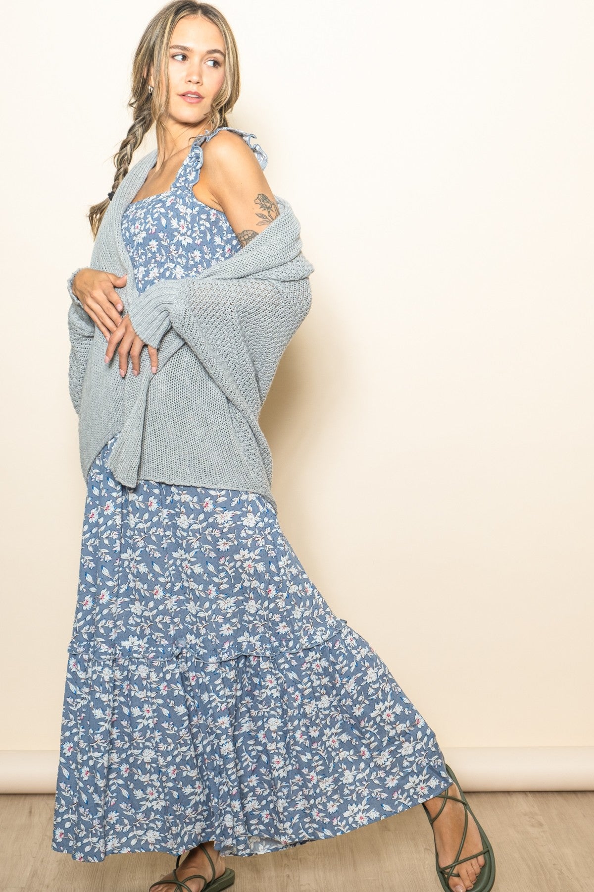 woman wearing a blue floral maxi dress with a ruffled hem and a light gray knit cardigan, striking a playful pose