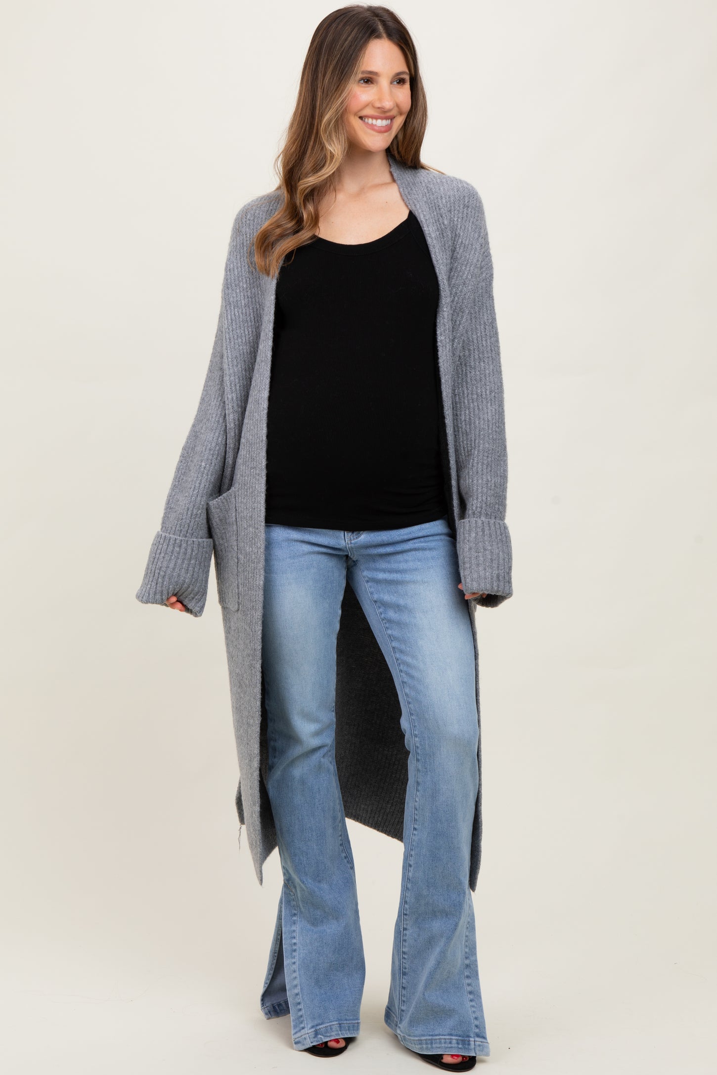 woman wearing a gray long cardigan over a black maternity top and blue flare jeans, showing her baby bump