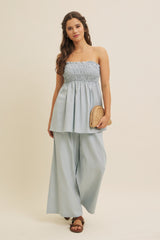woman wearing a light blue strapless smocked top with wide-leg pants, holding a straw clutch bag