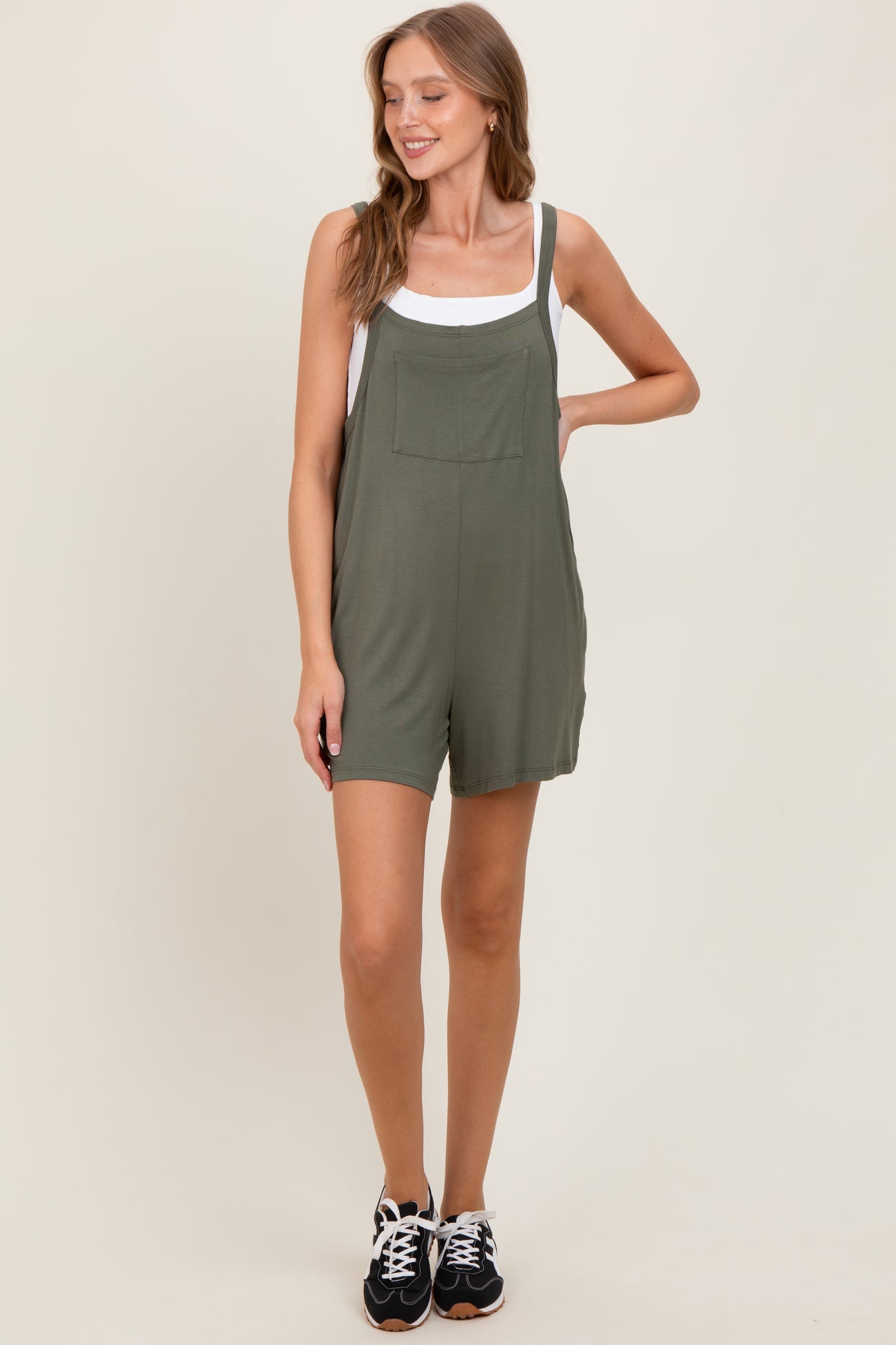 woman wearing an olive green short overalls with a white tank top underneath and black sneakers