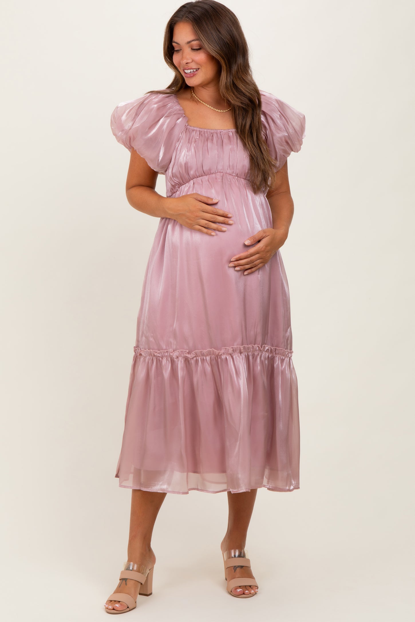 woman wearing a light pink satin maternity dress with puffy sleeves and a tiered skirt, holding her baby bump