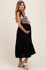 woman wearing a black maternity maxi dress with embroidered bodice and tie straps, holding her baby bump
