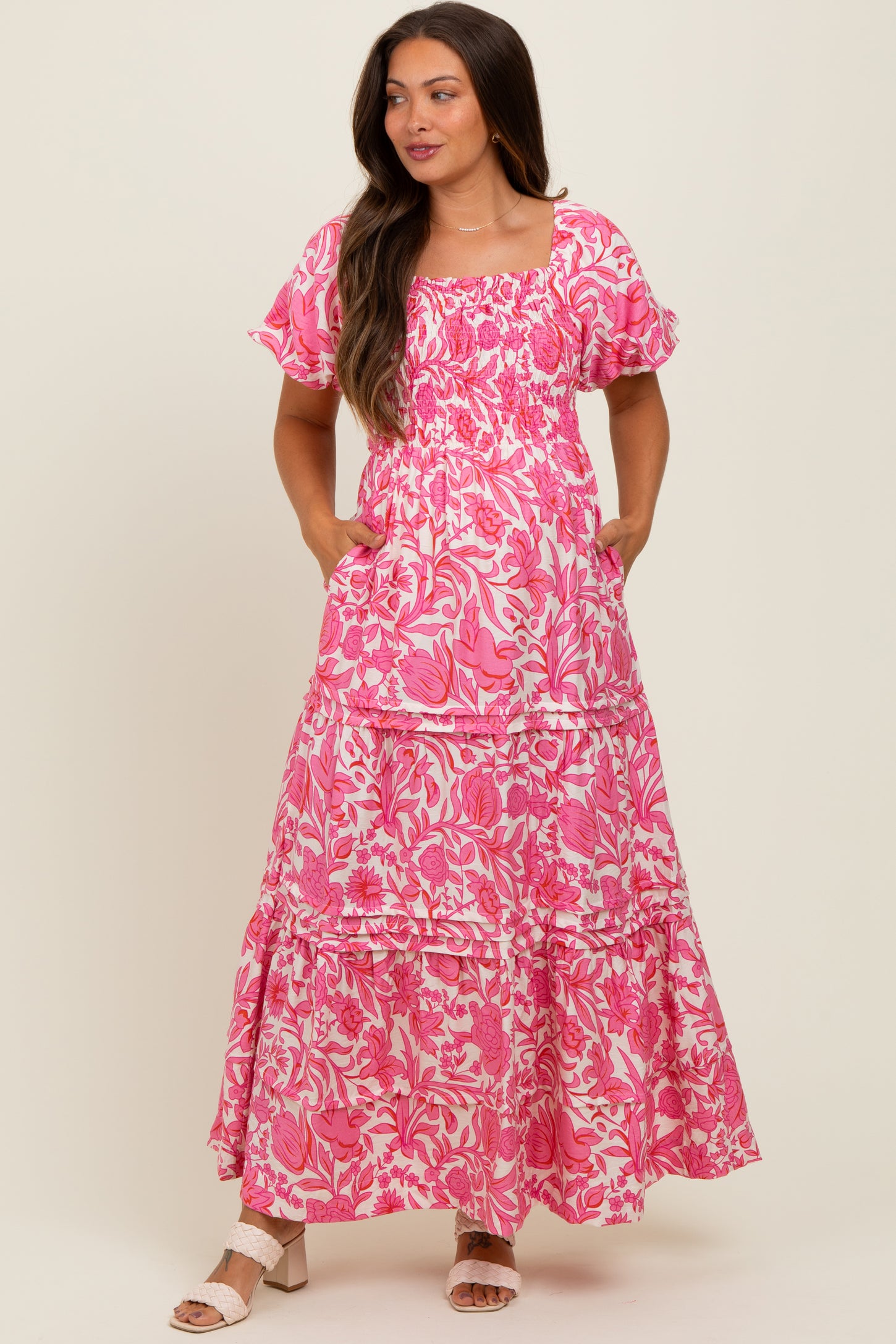 woman wearing a pink floral smocked maternity dress with short sleeves and pockets, featuring a tiered maxi design