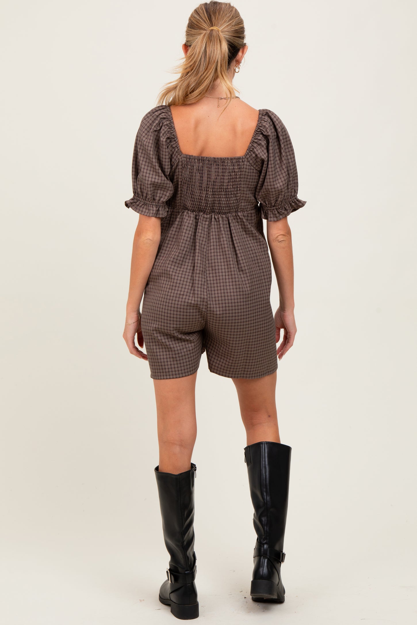 woman wearing a brown gingham maternity romper with puff sleeves, showing a smocked back and paired with knee-high black boots