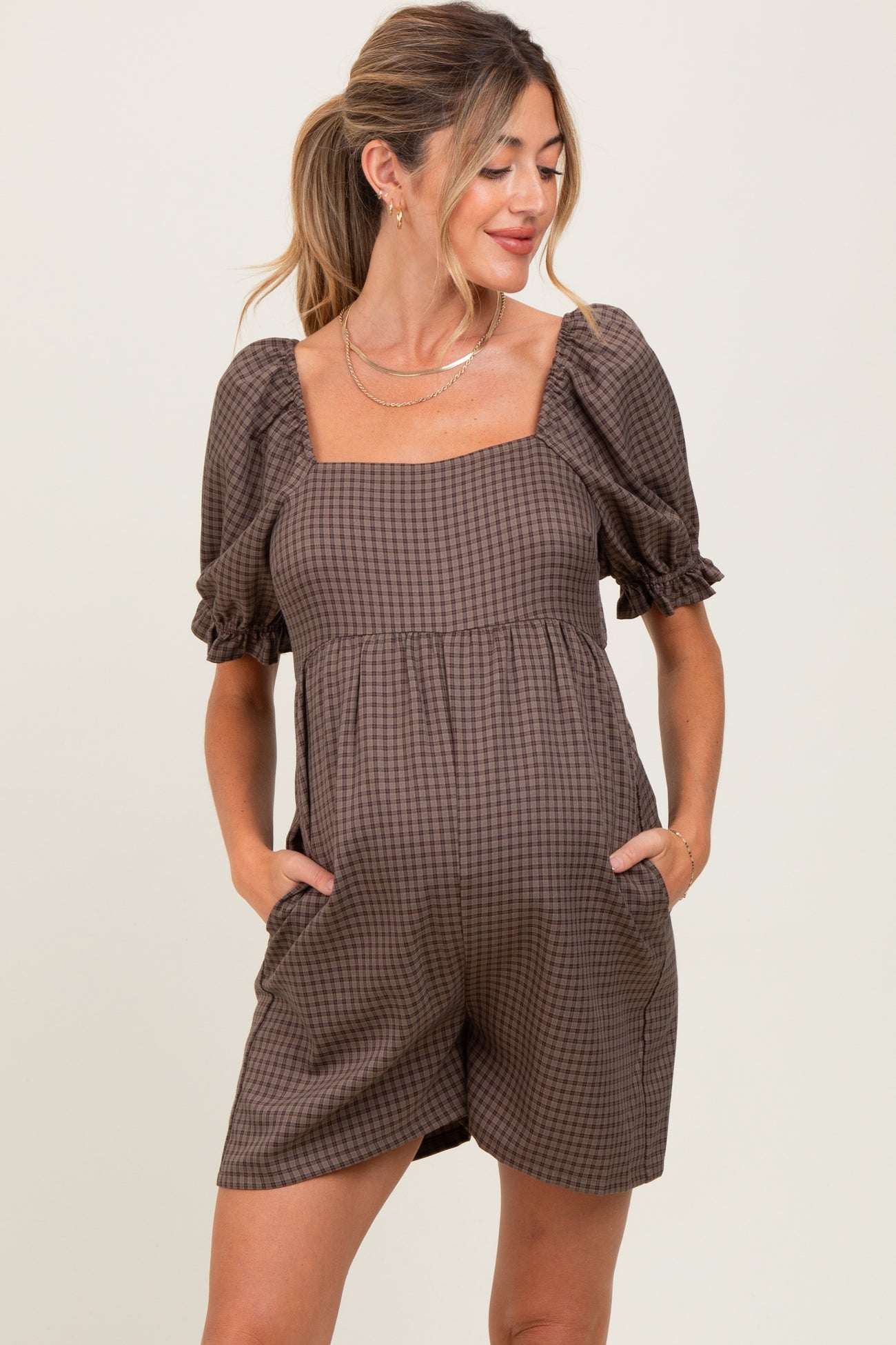 woman wearing a brown checkered maternity romper with short puff sleeves and pockets, showing her baby bump