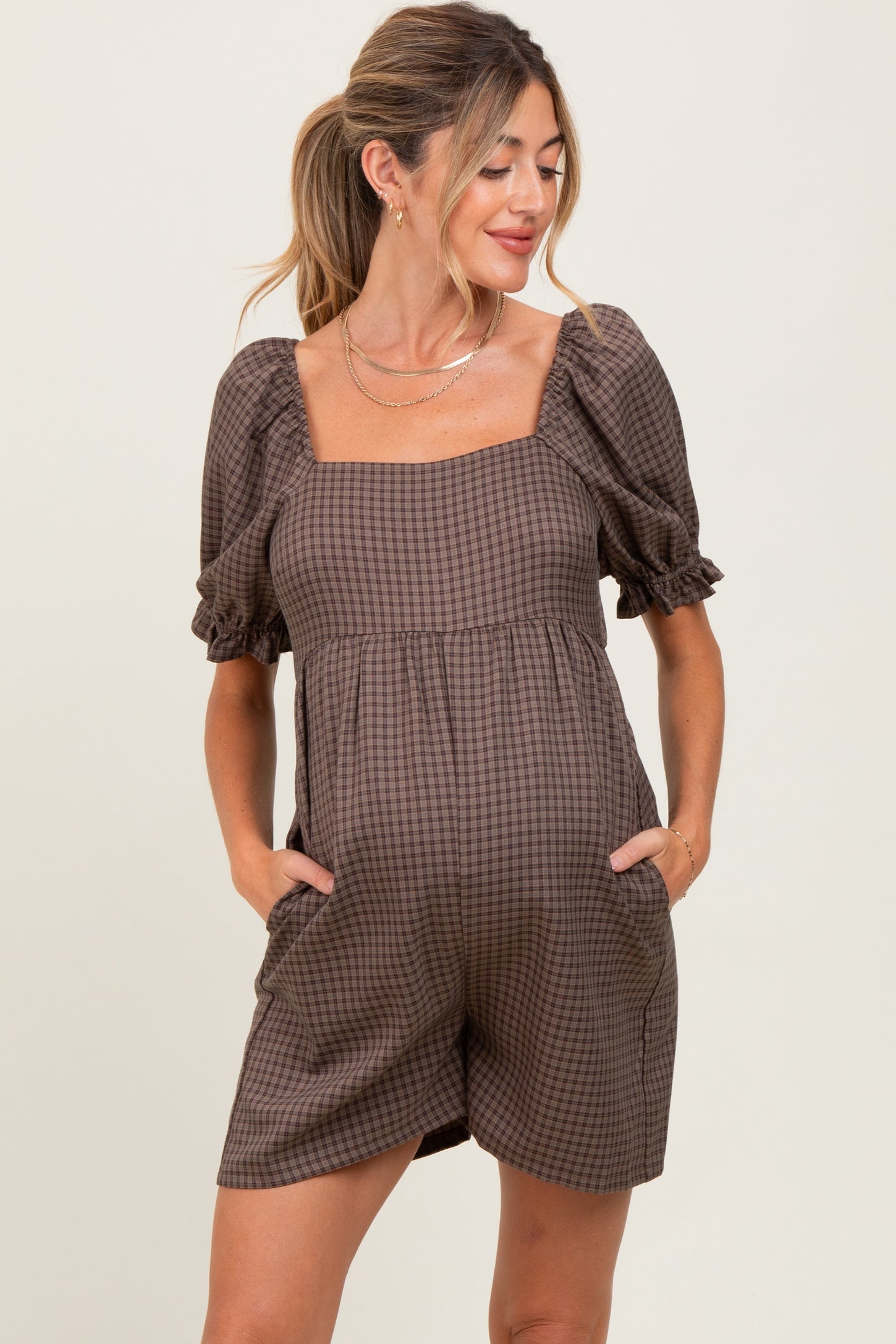 woman wearing a brown checkered maternity romper with short puff sleeves and pockets, showing her baby bump