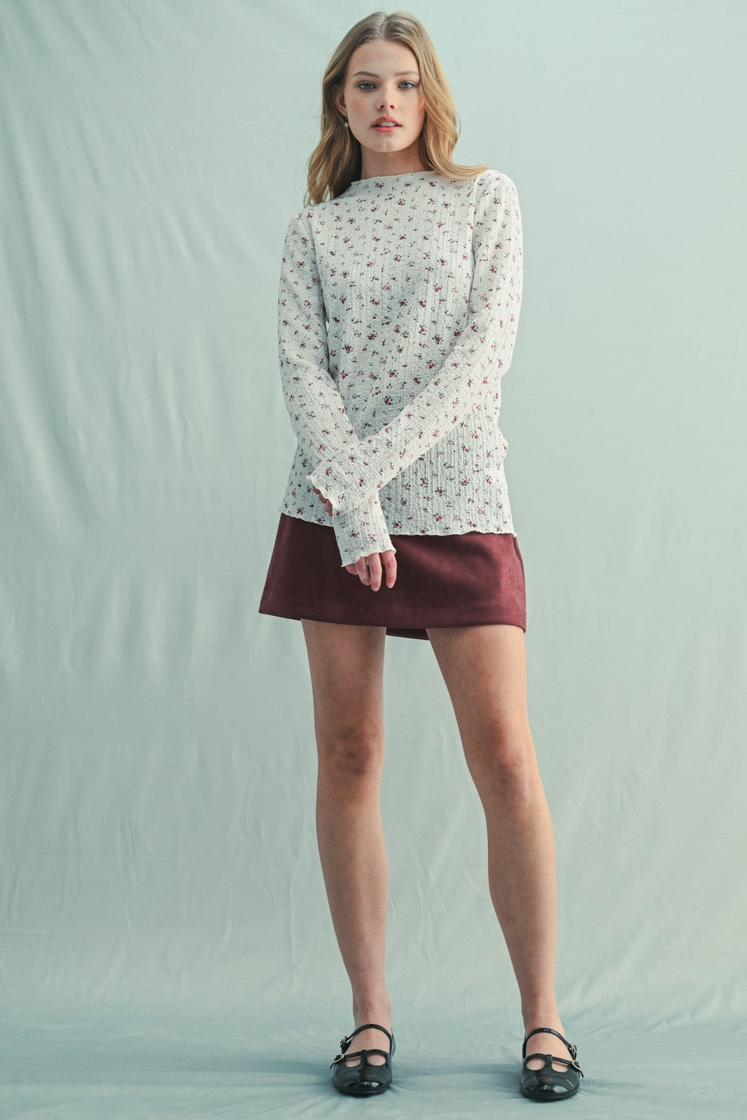 woman wearing a cream floral long sleeve top and a burgundy skirt, standing against a light background