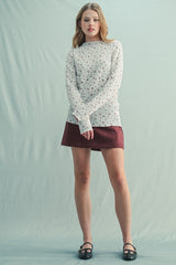 woman wearing a cream floral long sleeve top and a burgundy skirt, standing against a light background