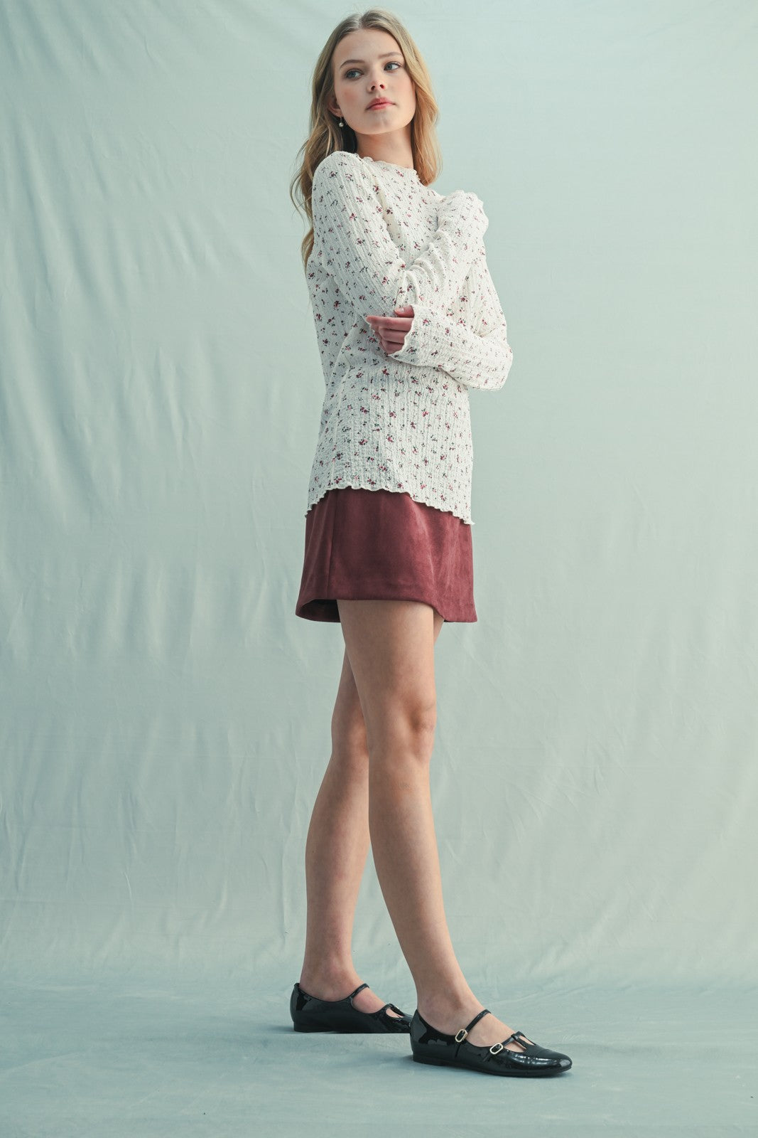 woman wearing a white crochet sweater over a maroon mini skirt and black flats, standing against a light backdrop