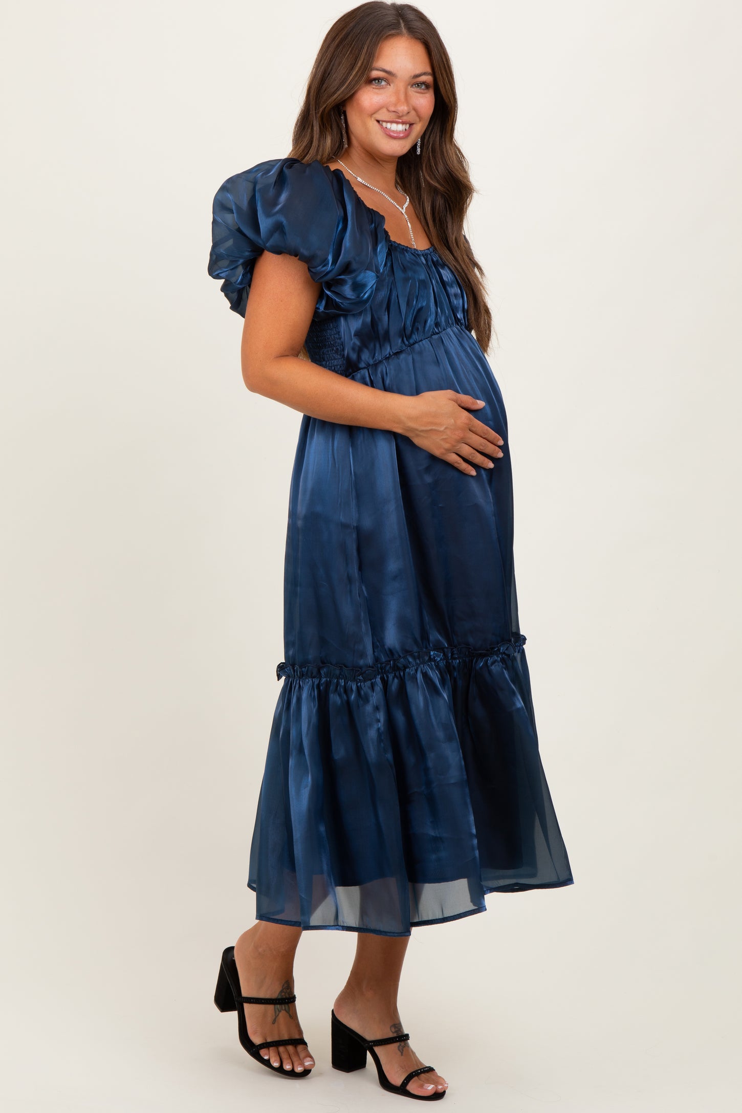 woman wearing a navy blue satin maternity dress with puff sleeves and a flowy skirt, cradling her baby bump