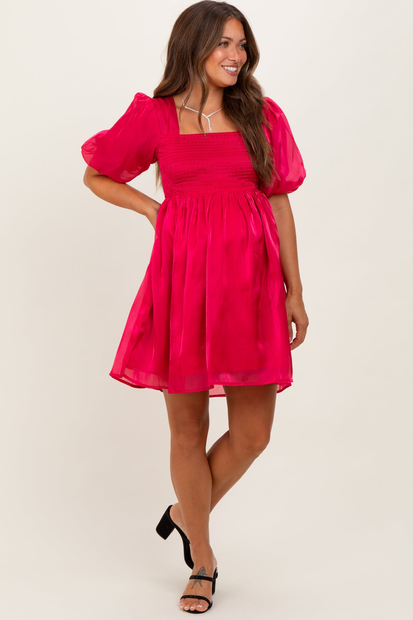 woman wearing a bright pink maternity dress with puff sleeves and a smocked bodice, standing and smiling